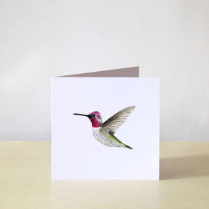 This note card features a hand-painted watercolor illustration of an Anna’s Hummingbird on a white background. The hummingbird is a soft brown colour with a bright pink neck and green under its wings. It’s depicted in flight. The greeting card is paired with a brown Kraft paper envelope.